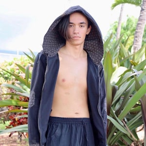 Puede incluir: Una persona con una chaqueta negra con capucha y un patrón geométrico y pantalones negros con un patrón geométrico. La persona está de pie frente a plantas verdes.