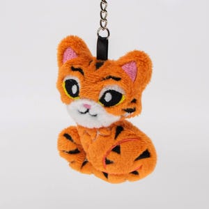 May include: A plush orange tiger keychain with black stripes, a white muzzle, and large black eyes. The tiger has pink inner ears and is attached to a metal chain with a black strap.