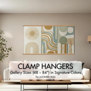 May include: A large, abstract art piece with a sunburst, arches, and geometric shapes in gold, blue, and green tones, displayed with wooden clamp hangers. The text reads "CLAMP HANGERS Gallery Sizes (48 - 84") in Signature Colors".