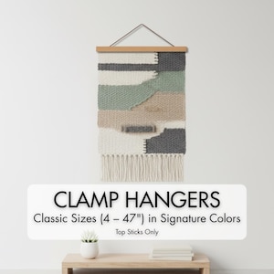 May include: A woven wall hanging with a wooden clamp and fringe. The textile features a geometric design in shades of white, gray, beige, and green. The text on the image reads "CLAMP HANGERS Classic Sizes (4 - 47") in Signature Colors".