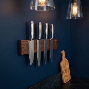 May include: Five stainless steel kitchen knives on a dark wood magnetic knife holder. A wooden cutting board rests against the wall. The background is a dark blue wall with two hanging lights.
