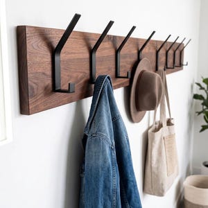 May include: A wooden wall-mounted coat rack with eight black metal hooks. A blue denim jacket, a brown hat, and a beige tote bag hang from the hooks. The rack is mounted on a white wall.