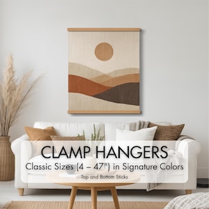 May include: A wall hanging with a landscape design in shades of brown, beige, and orange, displayed with wooden clamp hangers. The artwork features a sun and layered hills. The text on the image reads "CLAMP HANGERS Classic Sizes (4 - 47") in Signature Colors".
