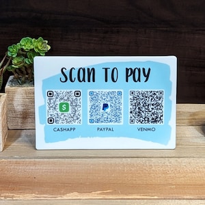 May include: A white sign with blue accents and the text "SCAN TO PAY" with three QR codes labeled "CASHAPP", "PAYPAL", and "VENMO".