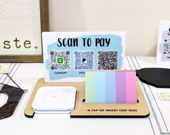 May include: A wooden stand with a white square card reader, a sign that says "Scan to Pay" with three QR codes for CashApp, PayPal, and an unknown app, and a stack of colorful sticky notes. The stand has a sign that says "Tap or Insert Chip Here".