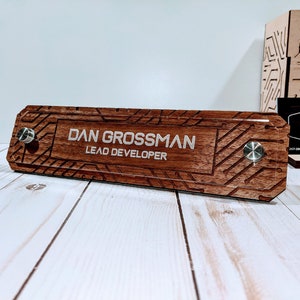 May include: A wooden desk nameplate with a silver engraved name "Dan Grossman" and the title "Lead Developer". The nameplate has a wood grain pattern and a circuit board design.