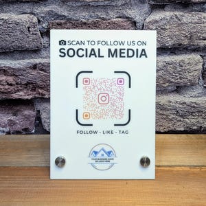 May include: White acrylic sign with the text "SCAN TO FOLLOW US ON SOCIAL MEDIA" and a QR code. Below the QR code are the words "FOLLOW . LIKE . TAG". The sign has a logo at the bottom and is mounted with two silver screws.
