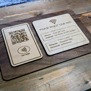 May include: Wooden WiFi sign with two sections. One section has a QR code and "TAP HERE" text. The other section says "PLEASE ENJOY OUR WIFI" with network and password details. The sign is on a wooden surface.