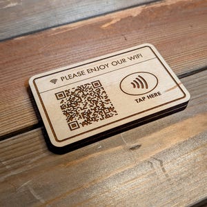 May include: Wooden rectangular sign with the text "PLEASE ENJOY OUR WIFI" and a QR code. The sign also features a Wi-Fi symbol and the words "TAP HERE". The sign is made of light-colored wood with dark brown engraved details.