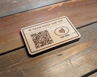 May include: Wooden rectangular sign with the text "PLEASE ENJOY OUR WIFI" and a QR code. The sign also features a Wi-Fi symbol and the words "TAP HERE". The sign is made of light-colored wood with dark brown engraved details.
