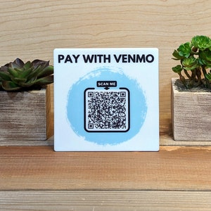 May include: A white square sign with blue and white circular design featuring a black and white QR code. The sign reads "PAY WITH VENMO" and "SCAN ME".