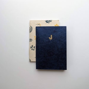 May include: Two notebooks, one with a floral pattern in yellow and green on a cream background, and the other a solid dark blue with a gold leaf design.