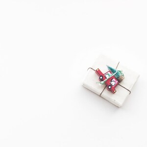 Puede incluir: Una caja de regalo blanca con un cami&oacute;n de juguete rojo y una peque&ntilde;a rama verde atada a la parte superior con cuerda.