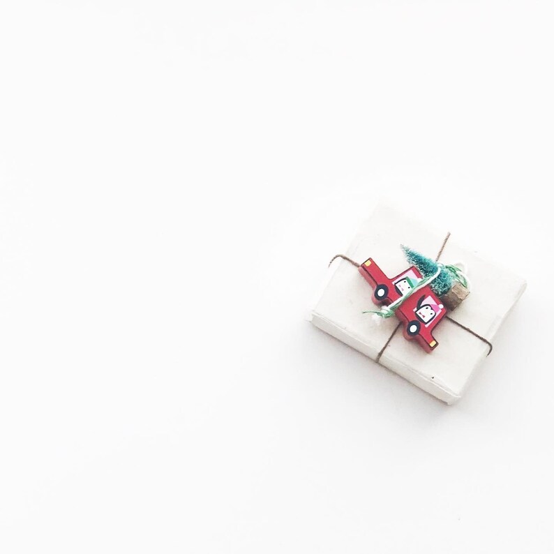Puede incluir: Una caja de regalo blanca con un cami&oacute;n de juguete rojo y una peque&ntilde;a rama verde atada a la parte superior con cuerda.