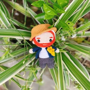 Könnte beinhalten: Eine gehäkelte Puppe mit einem braunen Hut, rotem Bart, blauer Jacke, weißem Hemd und grauer Hose. Die Puppe hält eine gelbe Blume und sitzt auf einer grünen Pflanze.