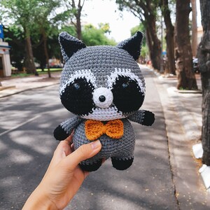 May include: A grey crocheted raccoon plush toy wearing an orange bow tie. The toy is being held in a hand.