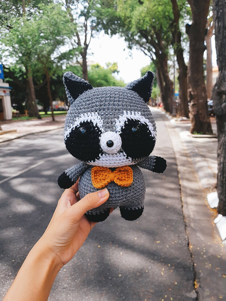May include: A grey crocheted raccoon plush toy wearing an orange bow tie. The toy is being held in a hand.