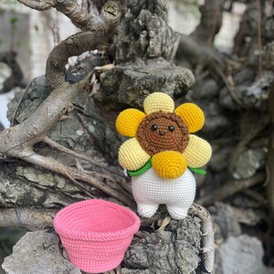 May include: A crocheted sunflower toy with a brown face and yellow petals. The toy is standing on a rock with a pink crocheted basket.