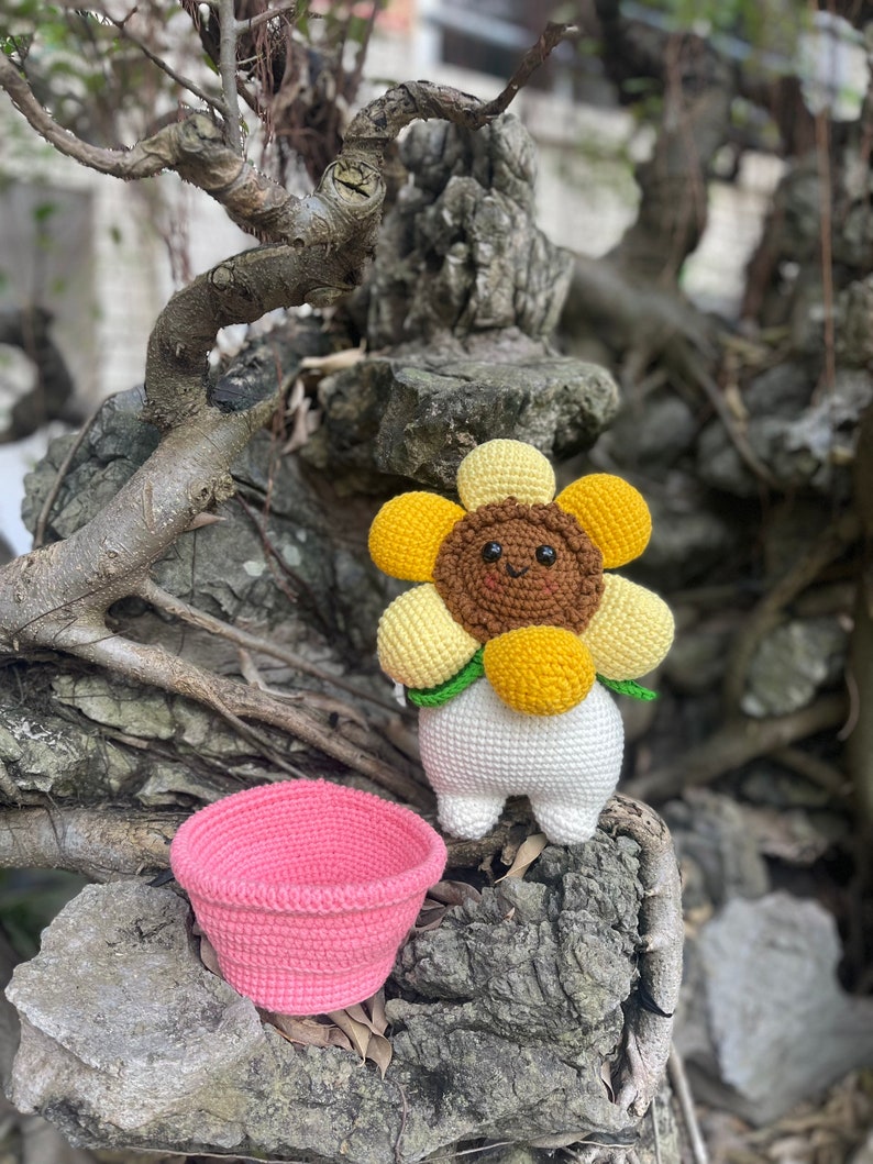 May include: A crocheted sunflower toy with a brown face and yellow petals. The toy is standing on a rock with a pink crocheted basket.