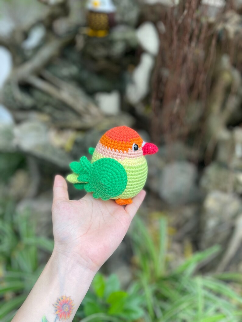 May include: A crocheted stuffed bird with green, orange, and yellow colors. The bird is sitting in a person's hand.