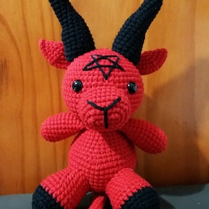 May include: A red crocheted plush toy shaped like a goat with black horns and hooves. The toy has a pentagram on its forehead and black button eyes.