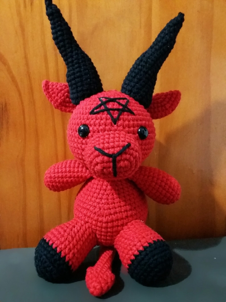 May include: A red crocheted plush toy shaped like a goat with black horns and hooves. The toy has a pentagram on its forehead and black button eyes.