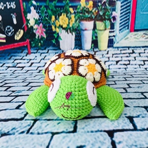 May include: A crocheted turtle toy with a brown and yellow shell decorated with white flowers. The turtle has green limbs and a white face with black eyes.