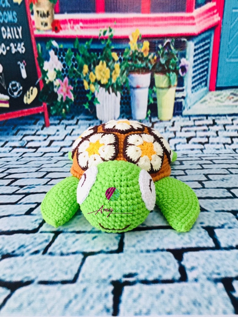 May include: A crocheted turtle toy with a brown and yellow shell decorated with white flowers. The turtle has green limbs and a white face with black eyes.