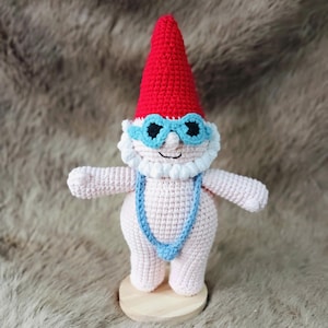 May include: A handmade crochet gnome doll with a red pointed hat, blue sunglasses, and a blue swimsuit. The gnome has a white beard and is standing on a wooden base. The doll is made of pink and white yarn.
