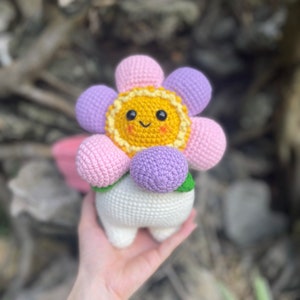 May include: A crocheted flower plush toy with a smiling face. The flower has pink, purple, and yellow petals and a white stem. The toy is being held in a person's hand.