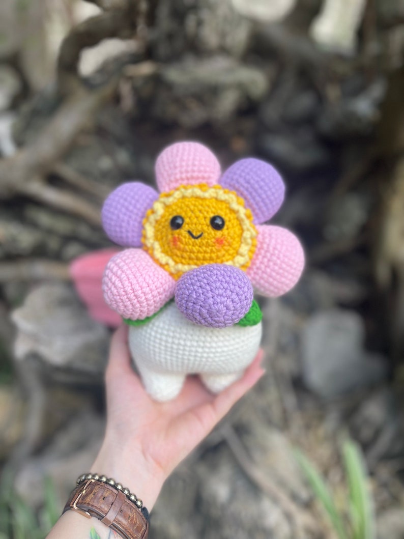 May include: A crocheted flower plush toy with a smiling face. The flower has pink, purple, and yellow petals and a white stem. The toy is being held in a person's hand.