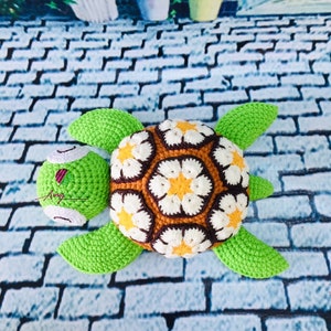 May include: A crocheted green turtle with a brown, white, and yellow flower pattern on its shell. The turtle is lying on a gray brick surface.