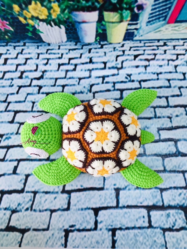 May include: A crocheted green turtle with a brown, white, and yellow flower pattern on its shell. The turtle is lying on a gray brick surface.