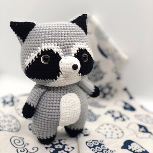 May include: A crocheted raccoon plush toy with black eyes, a black nose, and black and white markings on its face. The toy is standing on a white fabric with a blue pattern.