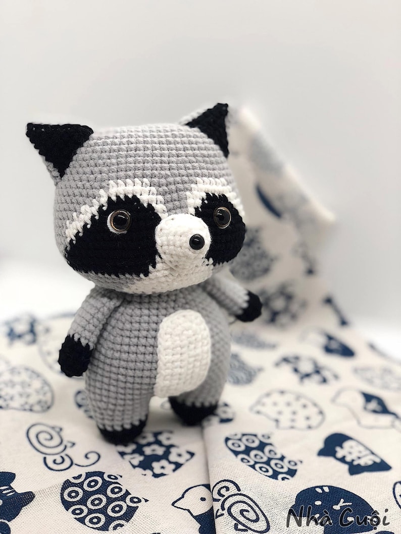 May include: A crocheted raccoon plush toy with black eyes, a black nose, and black and white markings on its face. The toy is standing on a white fabric with a blue pattern.