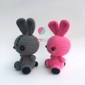May include: Two crocheted bunny plushies, one grey with red eyes and a stitched mouth, the other pink with black eyes and a stitched smile.