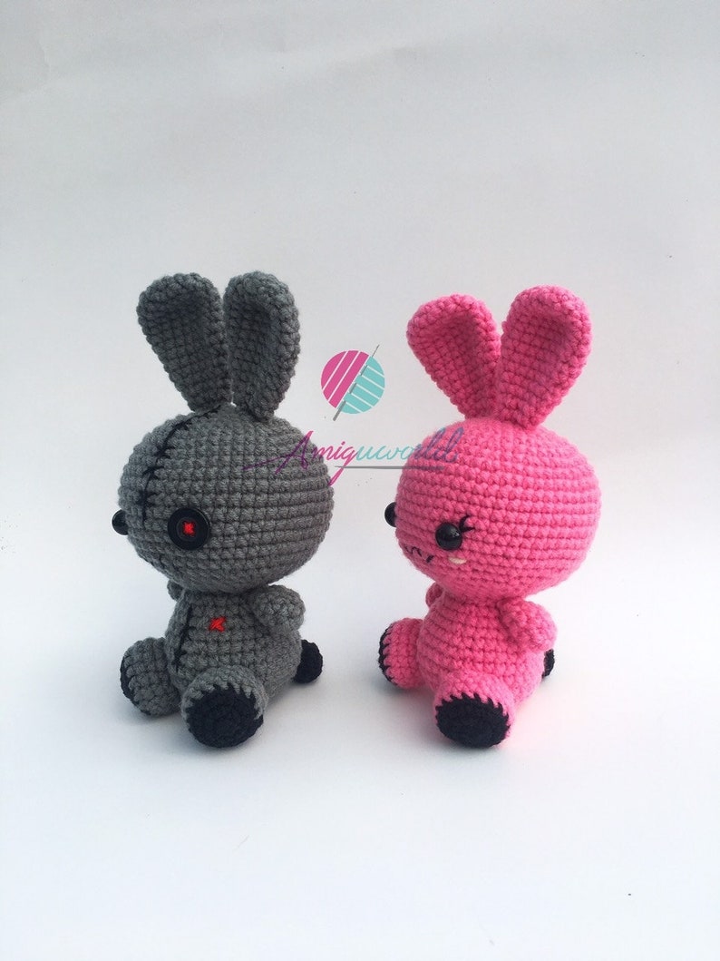 May include: Two crocheted bunny plushies, one grey with red eyes and a stitched mouth, the other pink with black eyes and a stitched smile.