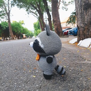 May include: A grey crocheted raccoon plush toy with black eyes and a black nose. The toy is standing on a grey asphalt road with a blurred background of trees and cars.
