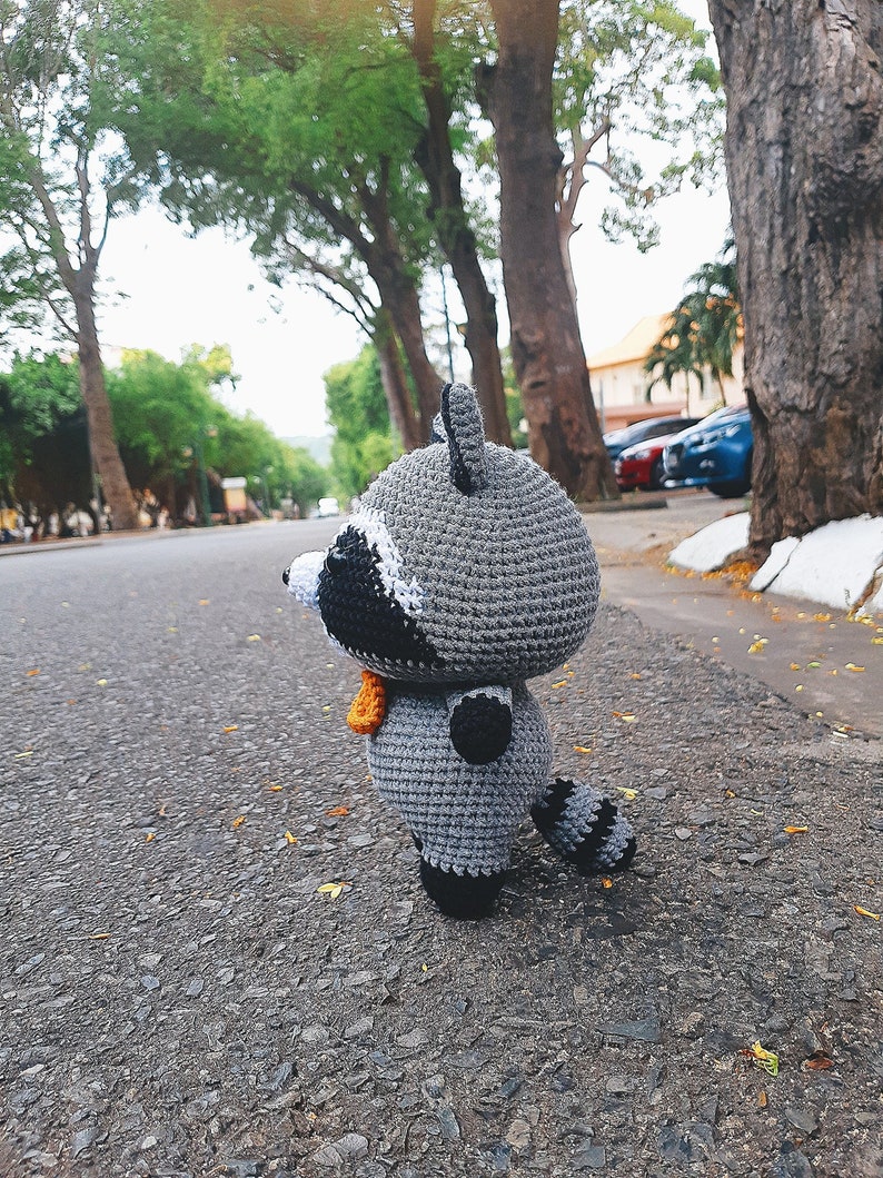 May include: A grey crocheted raccoon plush toy with black eyes and a black nose. The toy is standing on a grey asphalt road with a blurred background of trees and cars.