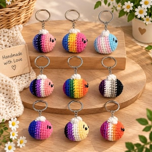 May include: Nine crochet bee keychains arranged in three rows on a wooden surface. Each bee features a unique color pattern, a white crocheted top, and a metal keyring. A small wooden sign with the text Handmade with Love is visible on the left.