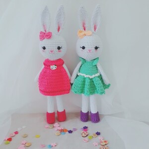 May include: Two white crocheted bunny dolls wearing dresses. One bunny is wearing a pink dress with a white button and the other bunny is wearing a green dress with a white button. Both bunnies have white bows in their hair.