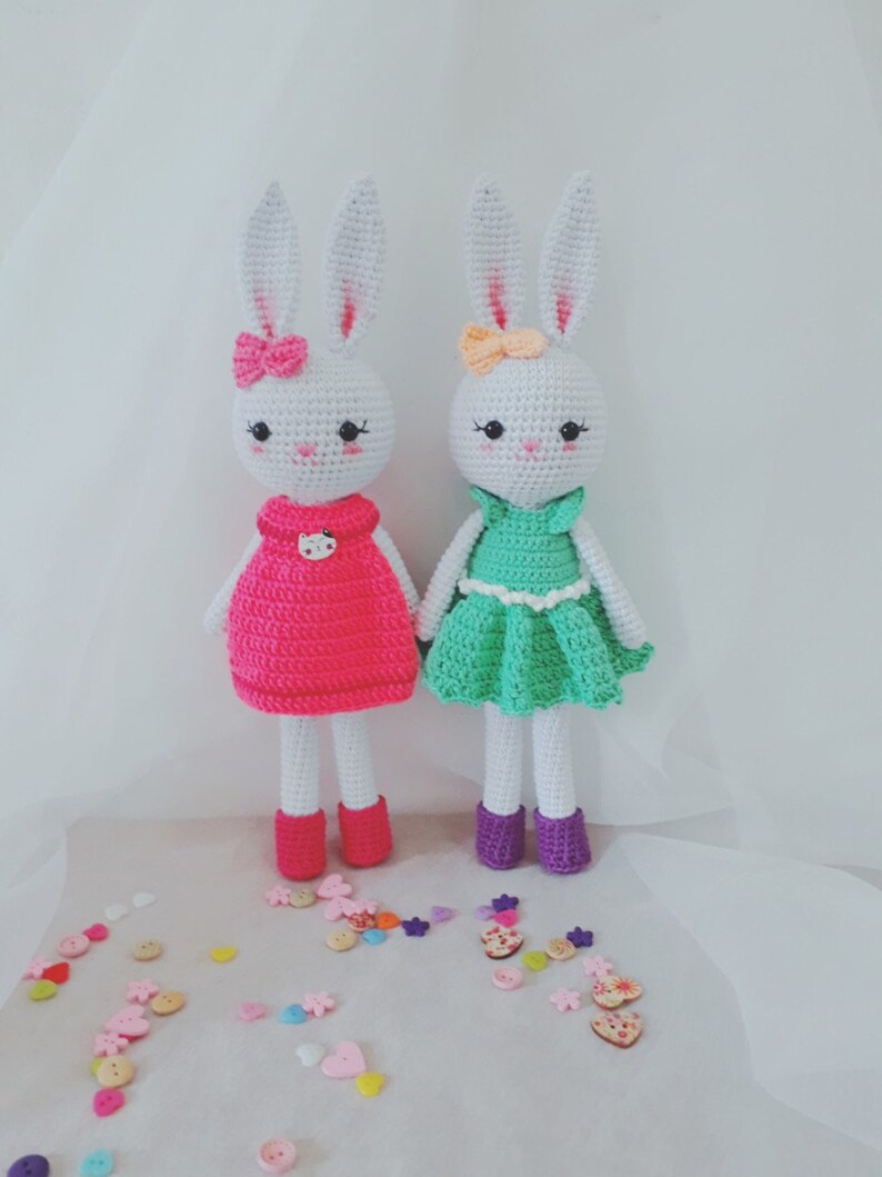 May include: Two white crocheted bunny dolls wearing dresses. One bunny is wearing a pink dress with a white button and the other bunny is wearing a green dress with a white button. Both bunnies have white bows in their hair.