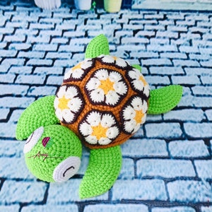 May include: A crocheted turtle toy with a green shell and brown and white flower pattern. The turtle has a green head and feet.