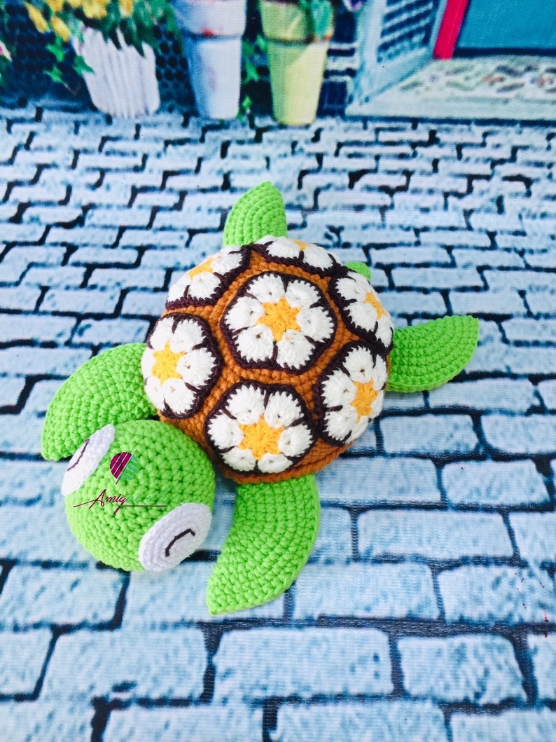 May include: A crocheted turtle toy with a green shell and brown and white flower pattern. The turtle has a green head and feet.