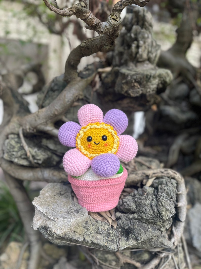 May include: A crocheted flower pot with a pink and purple flower and a yellow center. The flower has a smiling face.