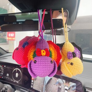 Könnte beinhalten: Vier gehäkelte Plüschtiere, die an einem Rückspiegel im Auto hängen. Die Spielzeuge sind ein lila und regenbogenfarbener Charakter, ein gelber Charakter mit einem schwarz-weißen Gesicht, ein rosa Charakter mit einem weißen Gesicht und ein lila Charakter mit einem weißen Gesicht.