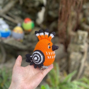 May include: A crocheted orange bird with black and white accents. The bird has a long beak and a crest of black and white feathers. The bird is being held in a person's hand.