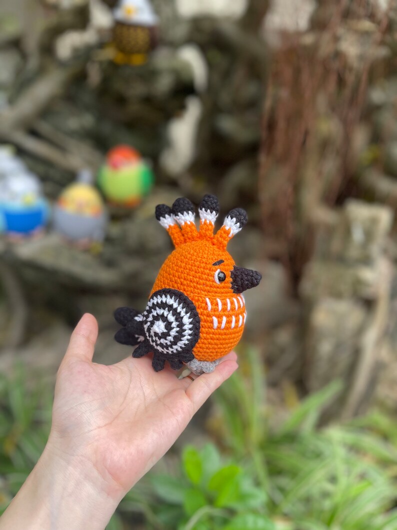 May include: A crocheted orange bird with black and white accents. The bird has a long beak and a crest of black and white feathers. The bird is being held in a person's hand.