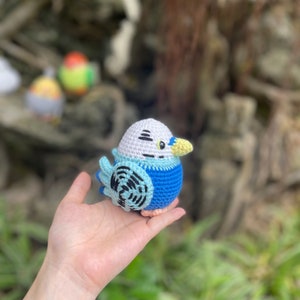 May include: A crocheted blue and white bird with a yellow beak and black eyes. The bird is sitting in a person's hand.