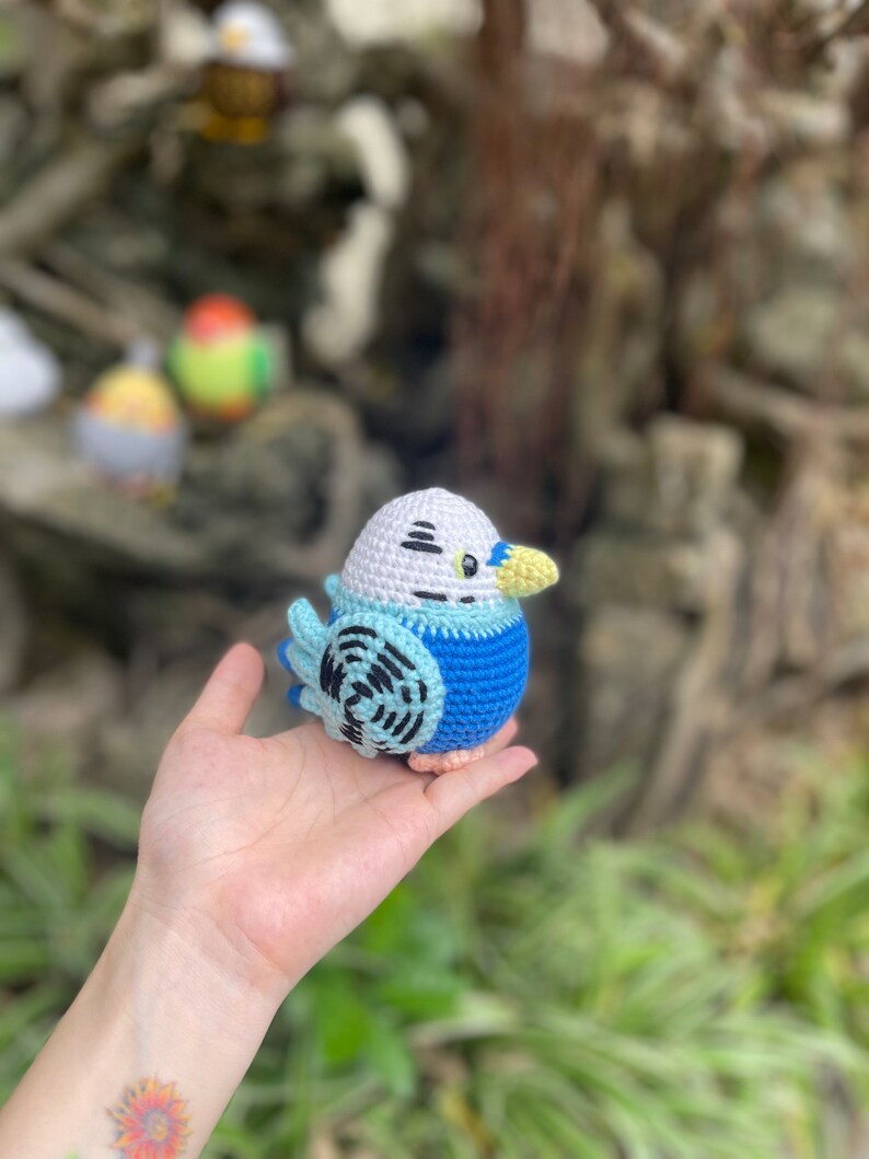 May include: A crocheted blue and white bird with a yellow beak and black eyes. The bird is sitting in a person's hand.
